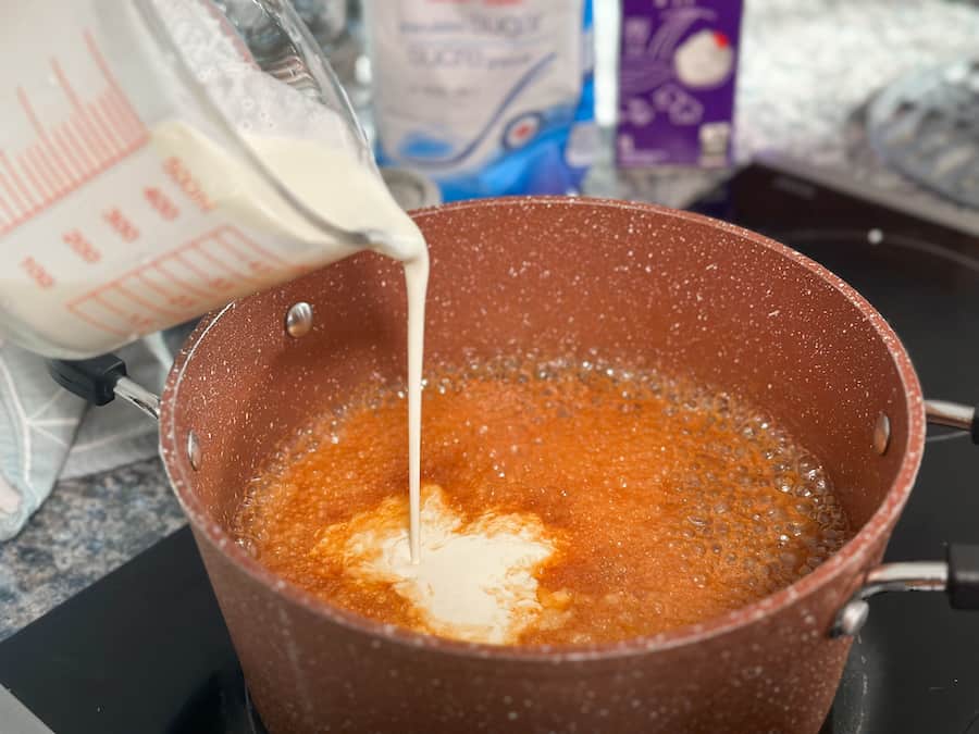 Slowly pouring whipping cream into the mixture of melted sugar and butter to make homemade salted caramel.