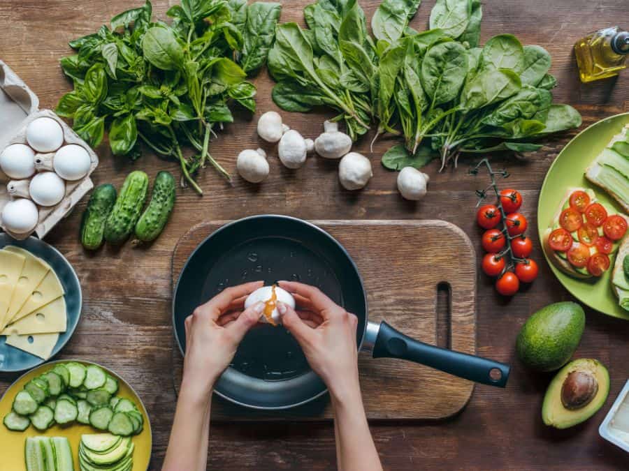 A women cracking an egg into a frying pan with vegetables all around the outside of the pan.