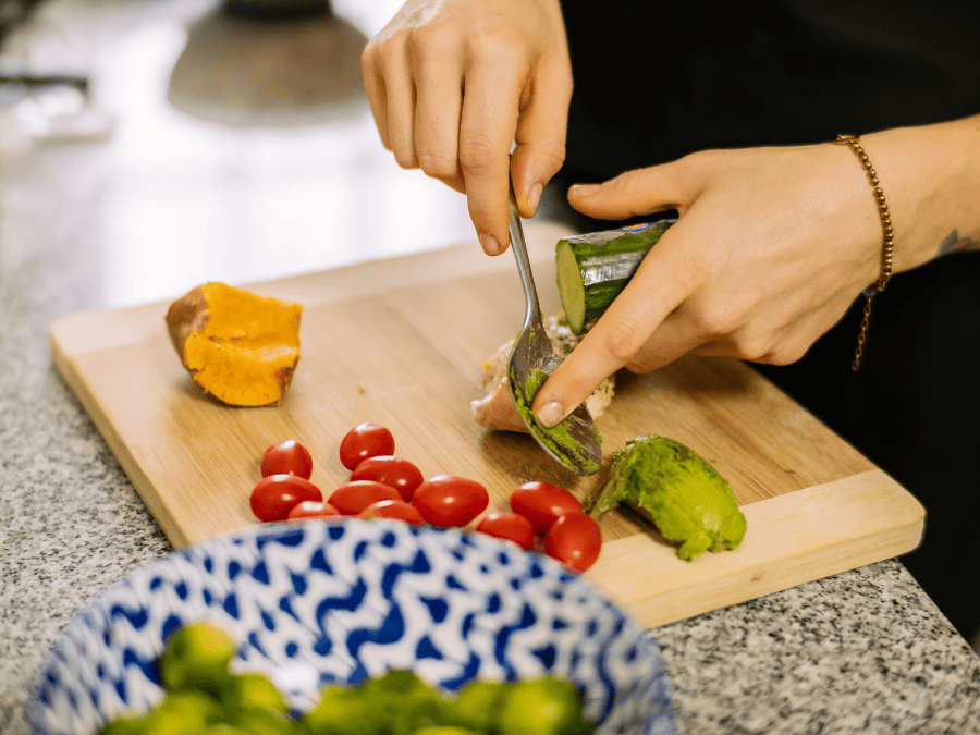 women with a spoon scrapping off avocado. There are tomatoes and cucumber also on a cutting board as she learns to cook from scratch. 