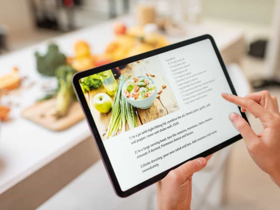 A tablet with a recipe on it that a women is about to use to learn to cook from scratch. In the background you can see vegetables that are blurred out.