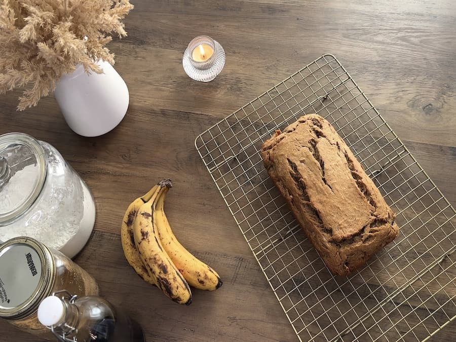 A loaf of gluten-free banana bread on a gold cooling rack and bananas off to the side. 