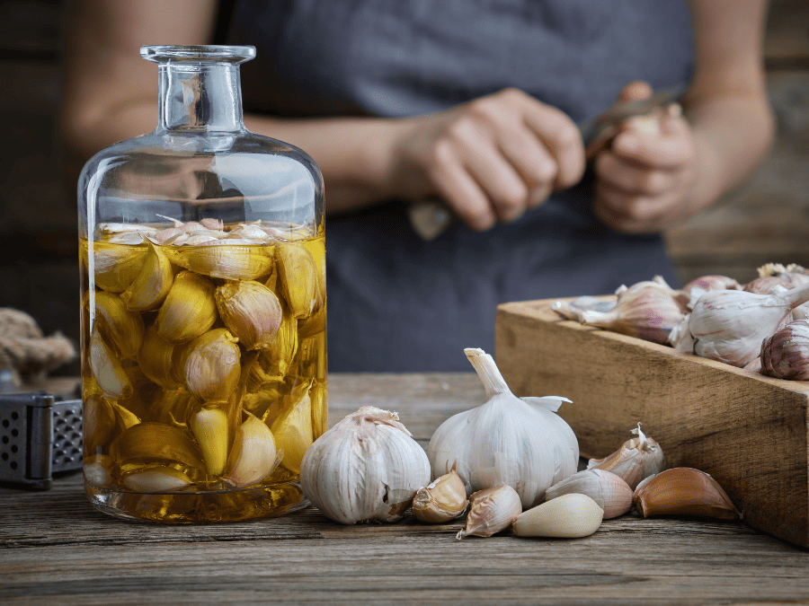 Garlic cloves in a glass jar of olive oil with a few garlic bulbs around the bottle. 