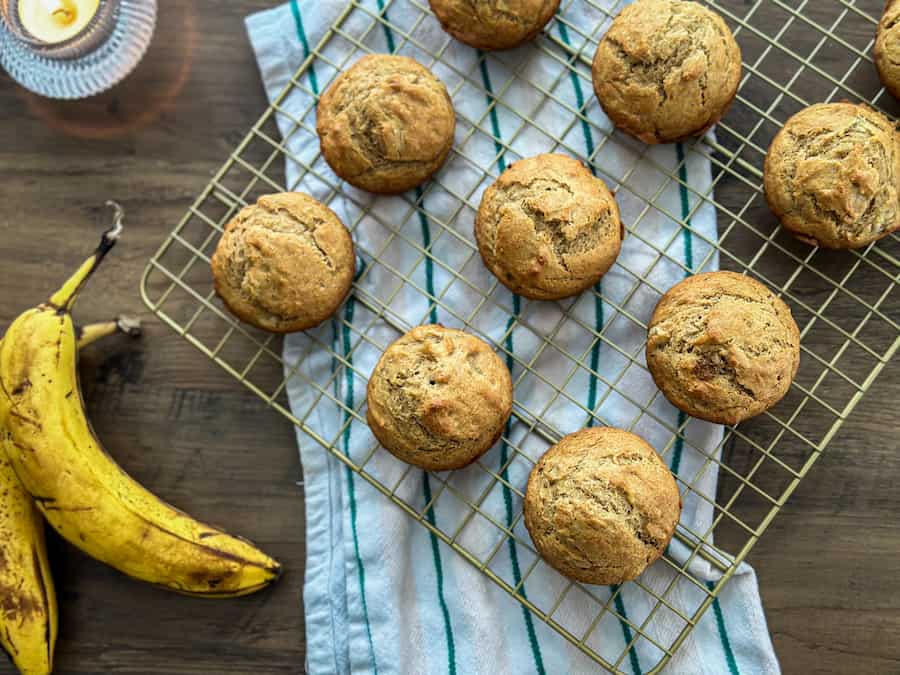 Gluten-Free Banana Bread Muffins on a gold cooling rack with a striped tea towel and two bananas beside it
