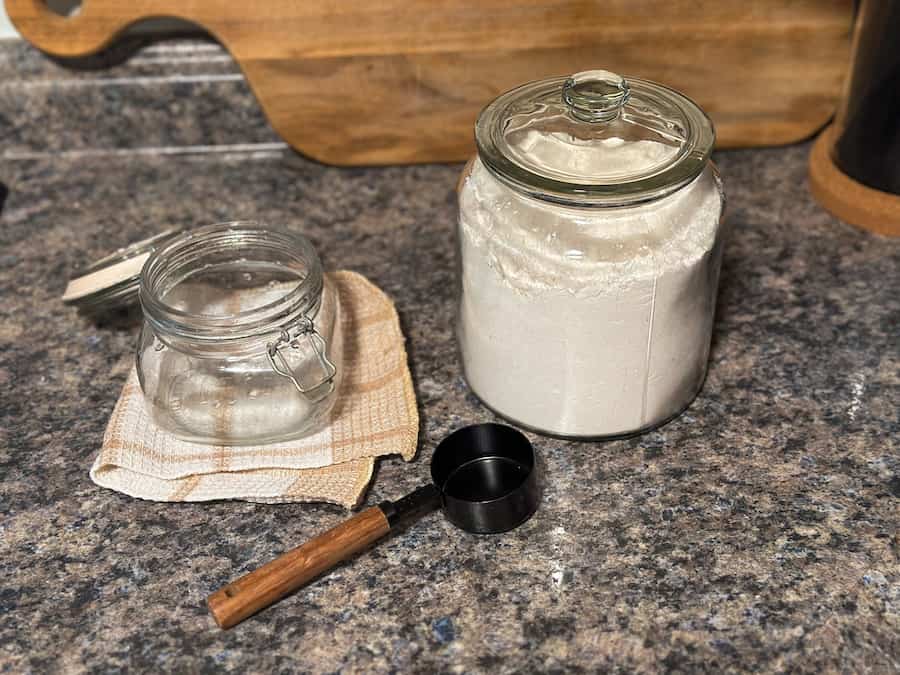 A glass jar, a jar of flour and a black measuring cup used to make sourdough starter. 