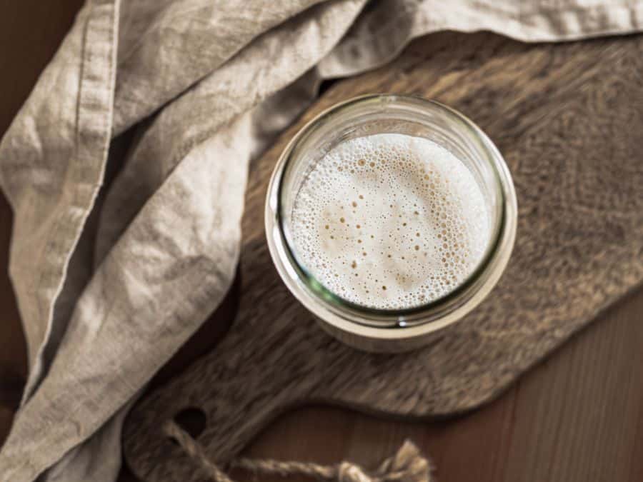 Sourdough starter in a glass jar on a wooden board with a linen cloth beside it. 